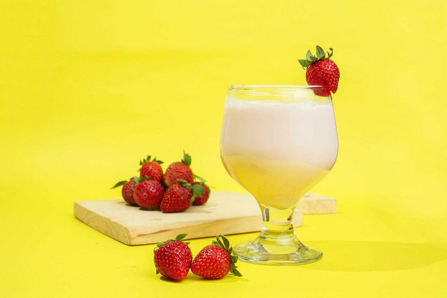 a glass of milk with strawberry garnish on cut board isolated yellow background photo