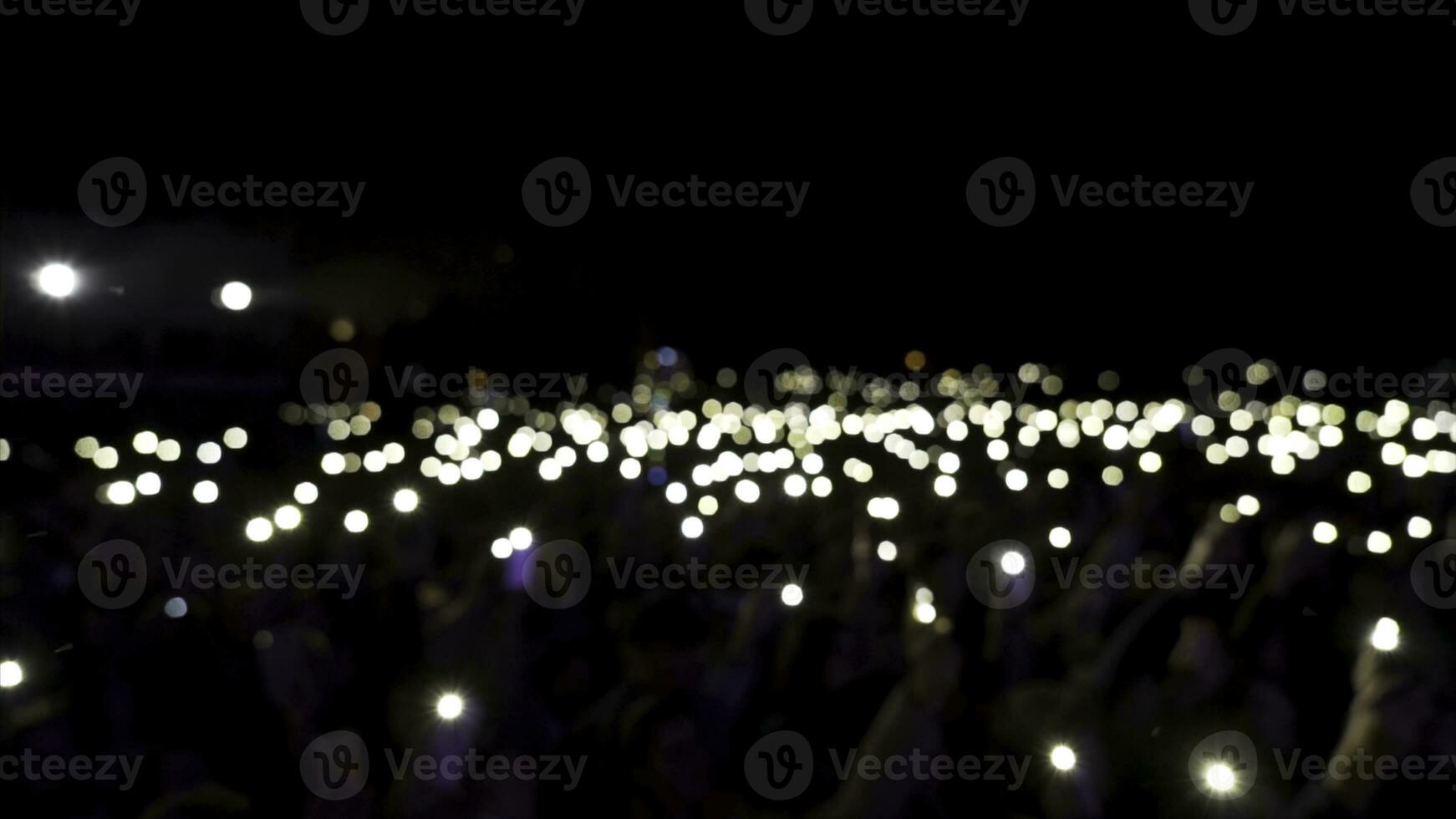 Defocused spectators in hall with lights waving in the dark. Action. Audience holding phones with shining flashlights during the concert, romantic atmosphere. photo