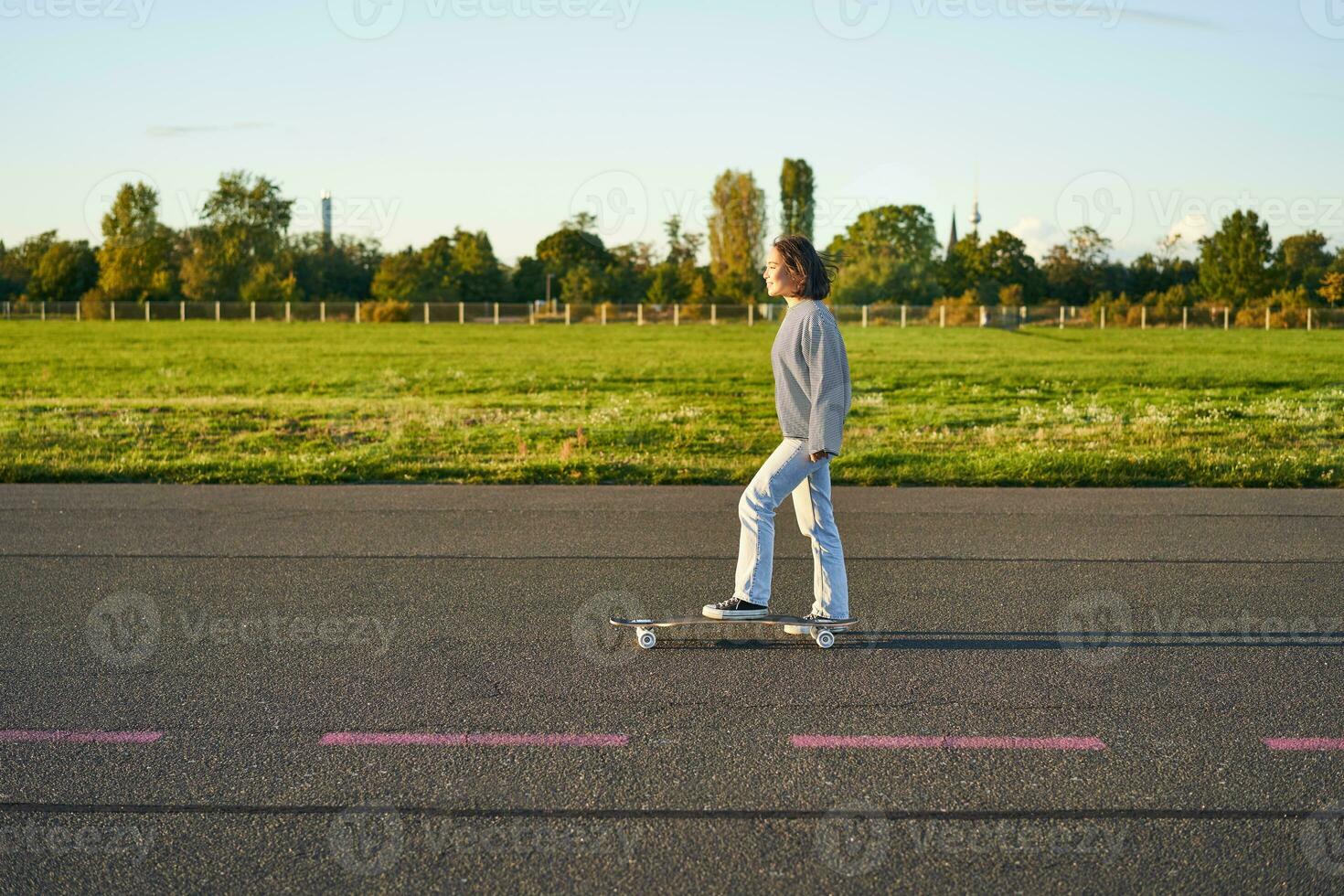 hermosa asiático patinador niña montando su longboard en soleado vacío la carretera. joven mujer disfrutando su patinar paseo sonriente y riendo foto