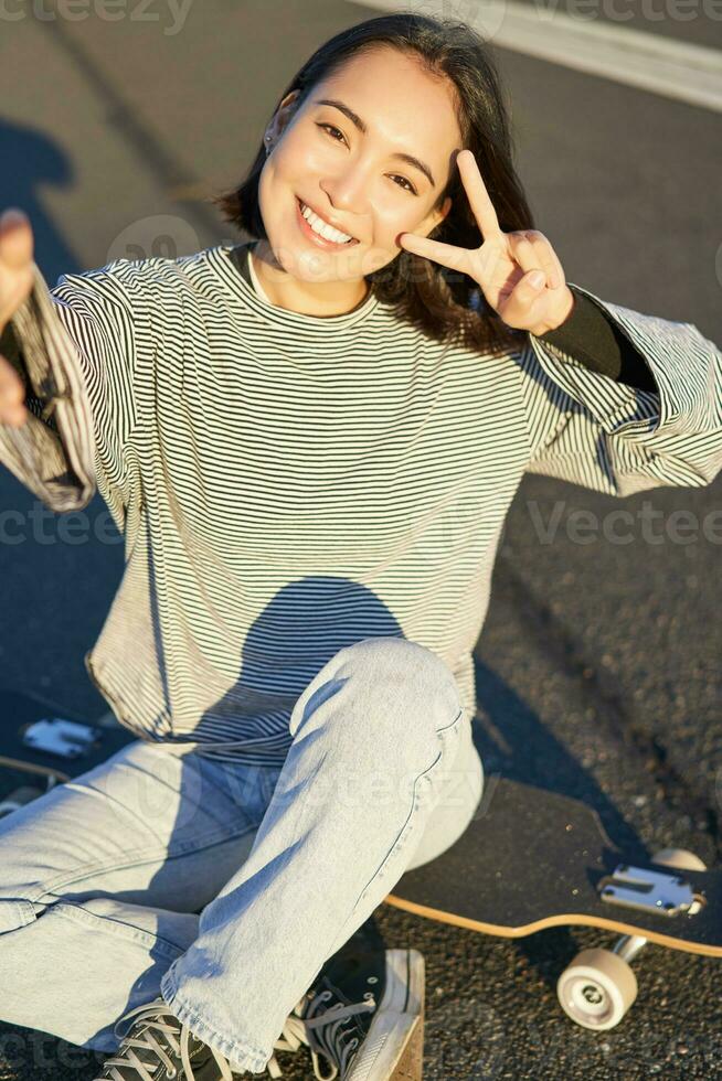 selfie de asiático niña sentado en patineta, tomando foto en teléfono inteligente, sonriente y demostración paz signo v