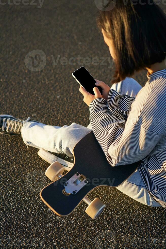 Vertical shot of asian girl typing, sending message or using mobile phone app, sitting on skateboard on road photo
