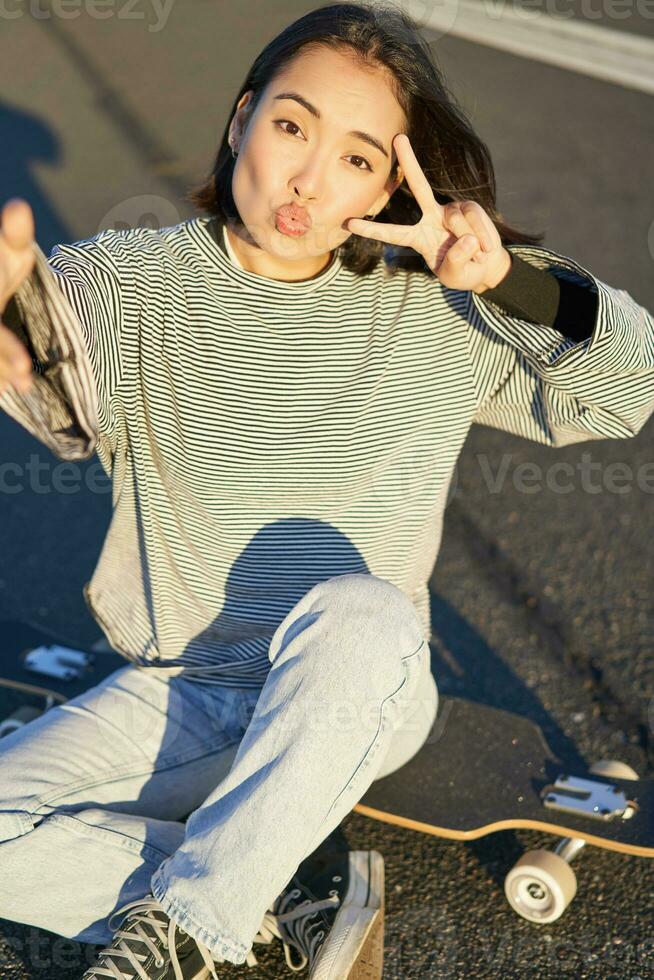 selfie de asiático niña sentado en patineta, tomando foto en teléfono inteligente, sonriente y demostración paz signo v