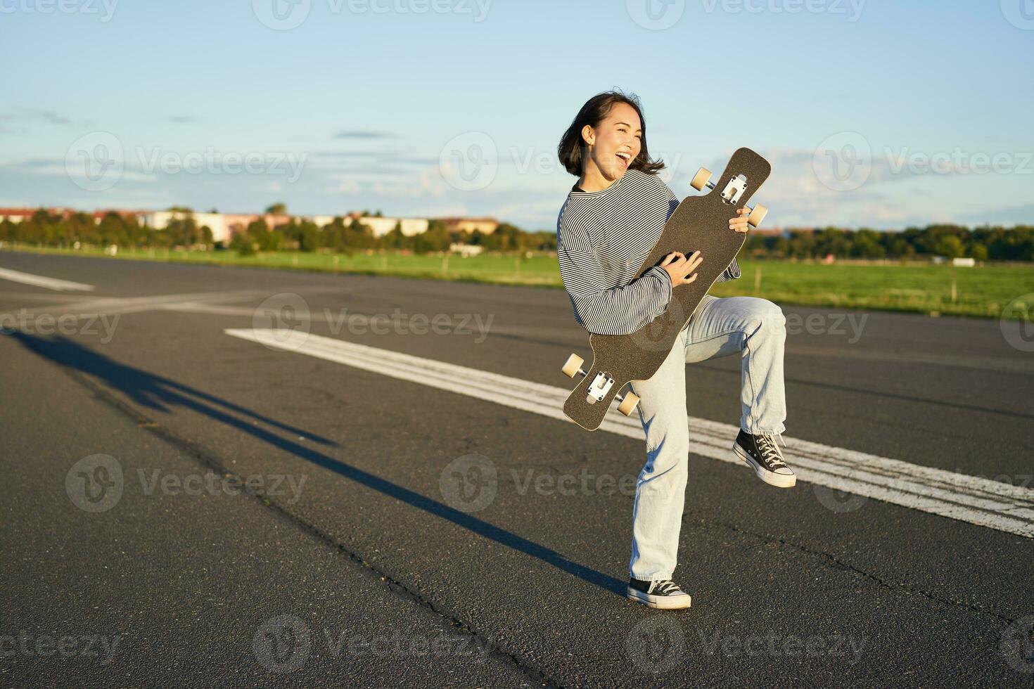 hermosa asiático adolescente niña jugando con su longboard, participación patineta como Si jugando guitarra, en pie en la carretera en soleado día foto