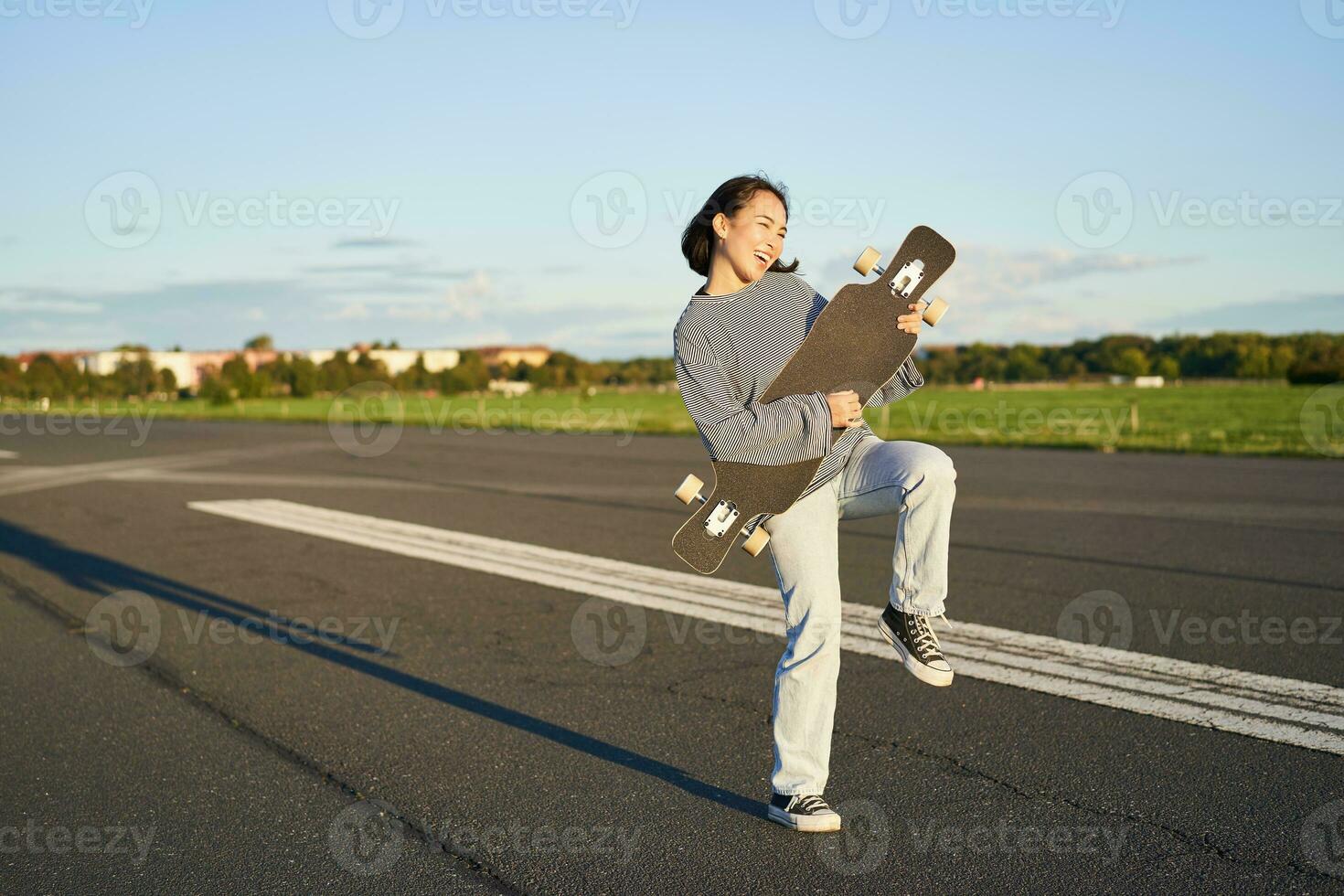 Happy brunette asian girl having fun on street with longboard, skating, using skateboard as guitar, enjoying skateboarding photo