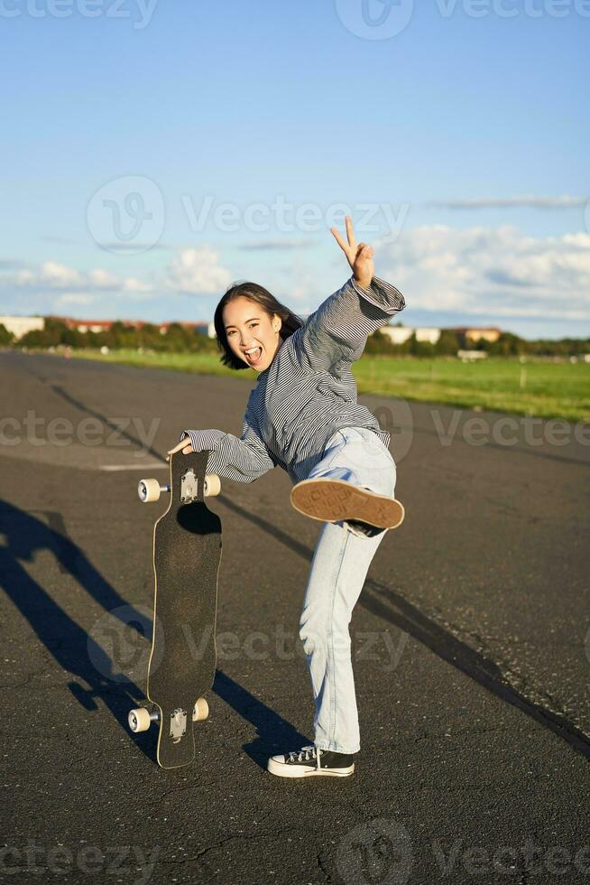 Vertical shot of happy asian skater girl, jumping, standing with skateboard and smiling. Woman skating on longboard and having fun photo