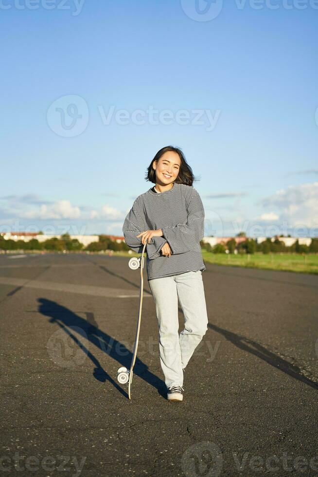 Vertical portrait of smiling asian woman standing on road with longboard, skateboarding on long cruiser, posing on empty road on sunny day photo