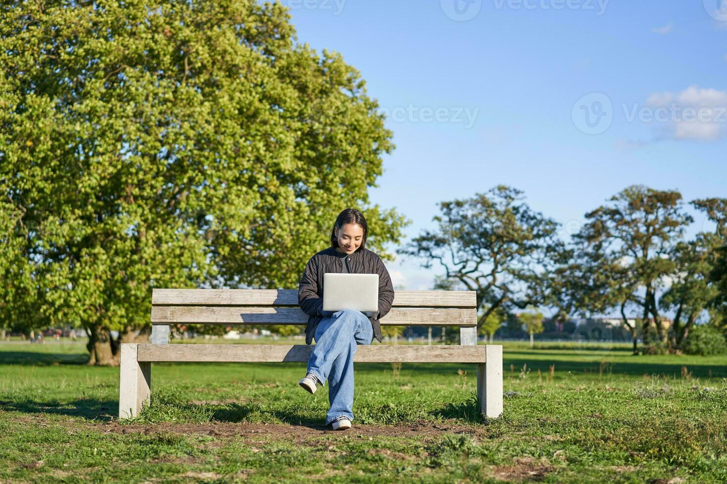 Portrait of young asian woman working on laptop, sitting on bench in park on sunny day, using her computer, studying in quite place photo