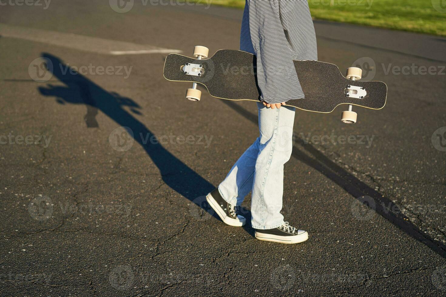 Cropped shot of teen girl body, holding cruiser longboard in hand, walking in sneakers on road in jeans and sweatshirt. Young woman skater with skateboard photo