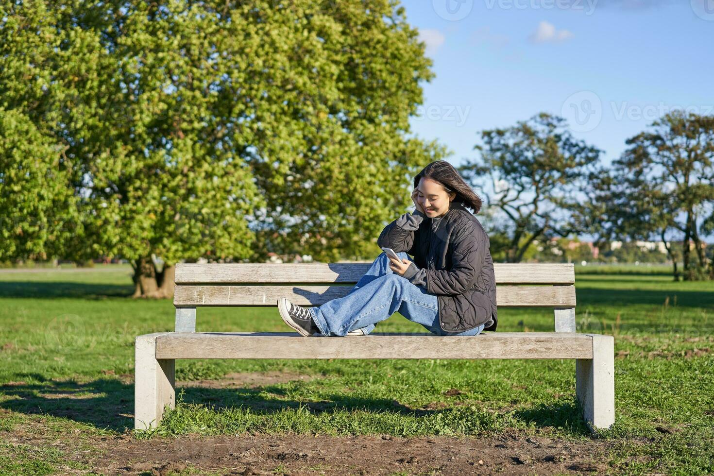 Young woman sitting alone on bench in park, using mobile phone, looking at screen, texting, sending messages in app chat photo