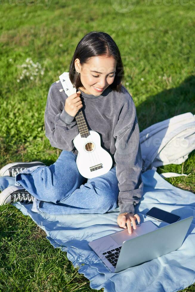 sonriente asiático niña aprende cómo a jugar ukelele en computadora portátil, vídeo charla con música maestro, sentado con instrumento en parque en césped foto