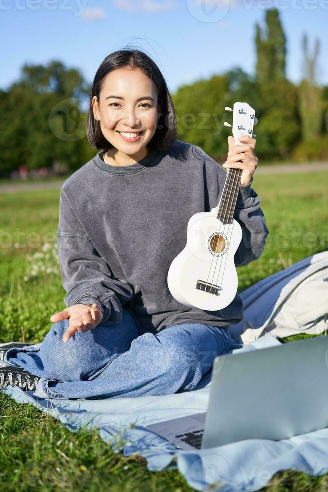 vertical Disparo de contento asiático niña obras de teatro instrumento, muestra su ukelele a ordenador portátil cámara, vídeo chats acerca de música, enseña cómo a jugar, se sienta en parque al aire libre foto