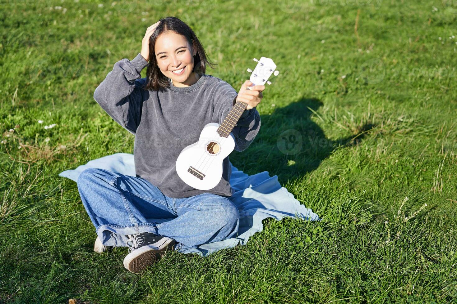 emocionado asiático niña se sienta en parque con ukelele, obras de teatro instrumento y siente contento optimista, positivo momentos y personas concepto foto