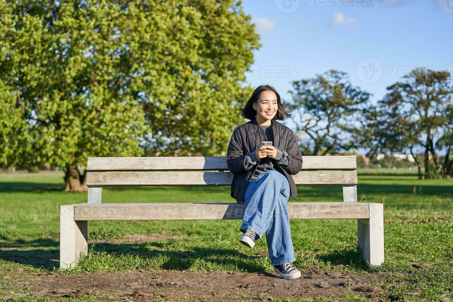 Young woman sitting alone on bench in park, using mobile phone, looking at screen, texting, sending messages in app chat photo