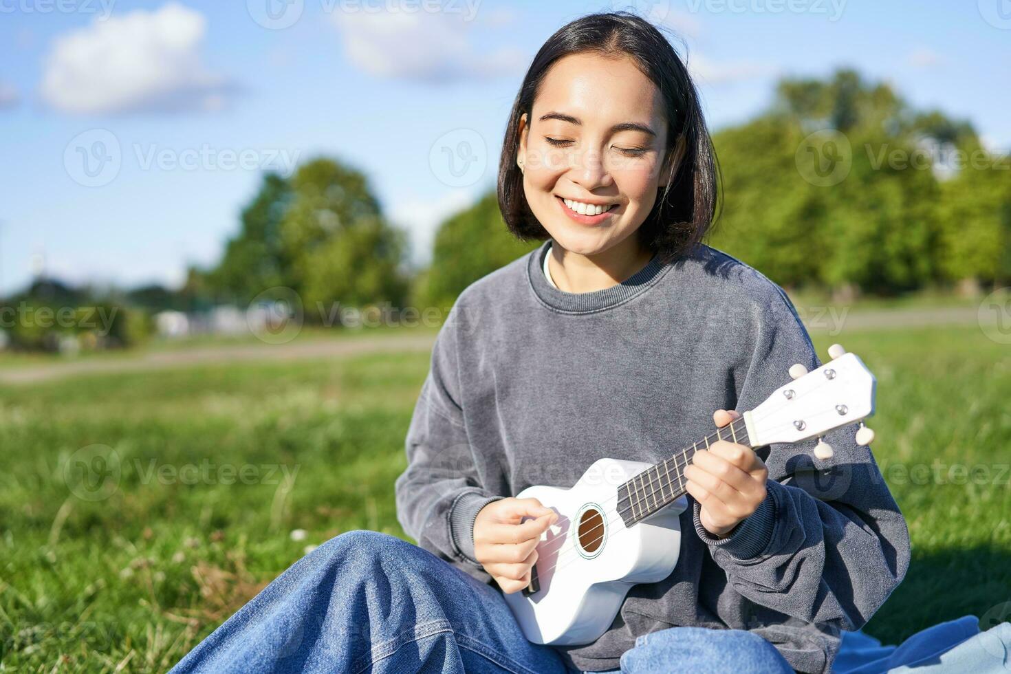 Carefree asian girl singing and playing ukulele in park, sitting on grass, musician relaxing on her free time outdoors photo