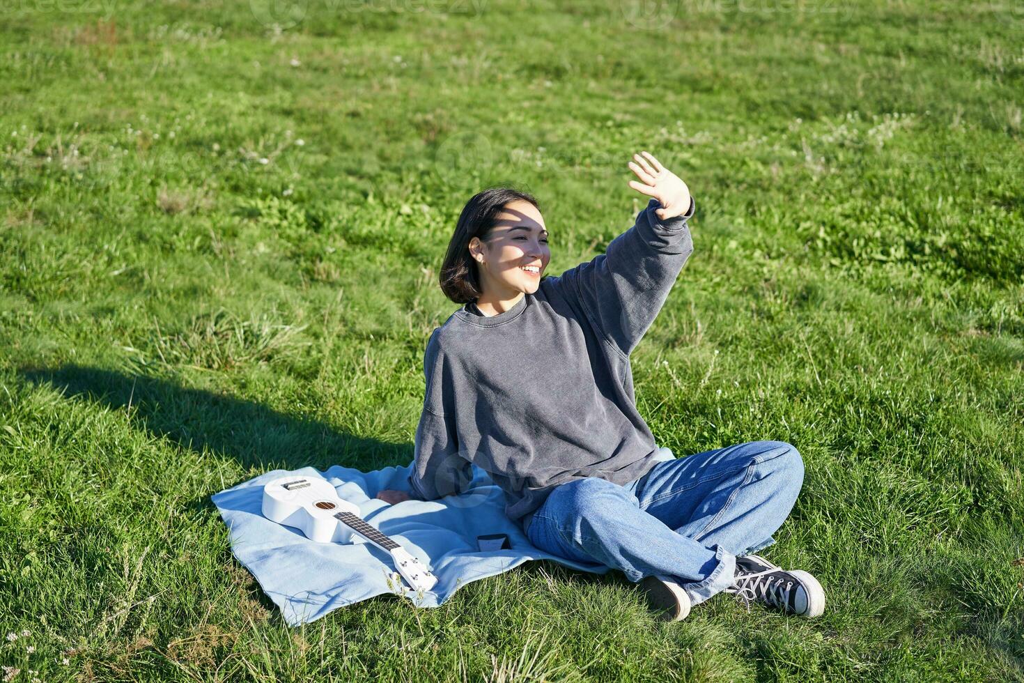 contento asiático niña sentado en picnic cobija con ukelele, cubrir sí misma desde luz de sol, extensión mano hacia Dom vigas y sonriente foto