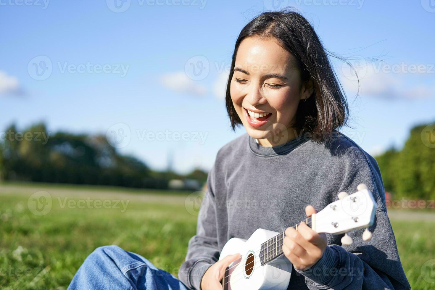 contento personas y aficiones. sonriente asiático niña jugando ukelele guitarra y cantando, sentado en parque al aire libre en cobija foto