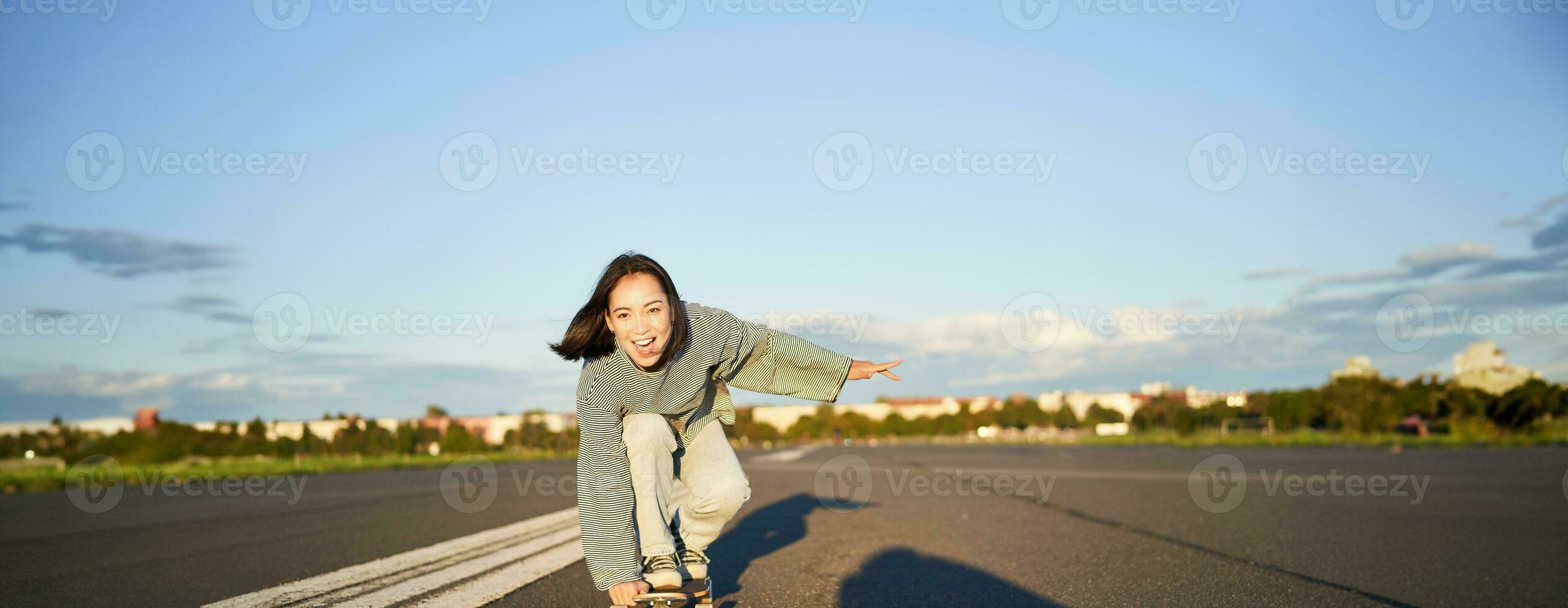 Skater girl riding on skateboard, standing on her longboard and laughing, riding cruiser on an empty street towards the sun photo