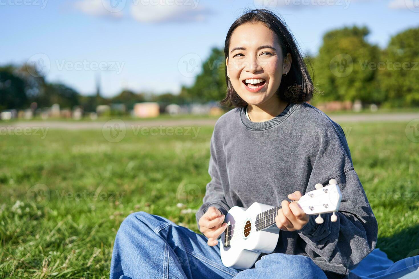 retrato de asiático niña alumno, jugando ukelele y canto en parque, sentado solo en cobija y disfrutando haciendo música foto