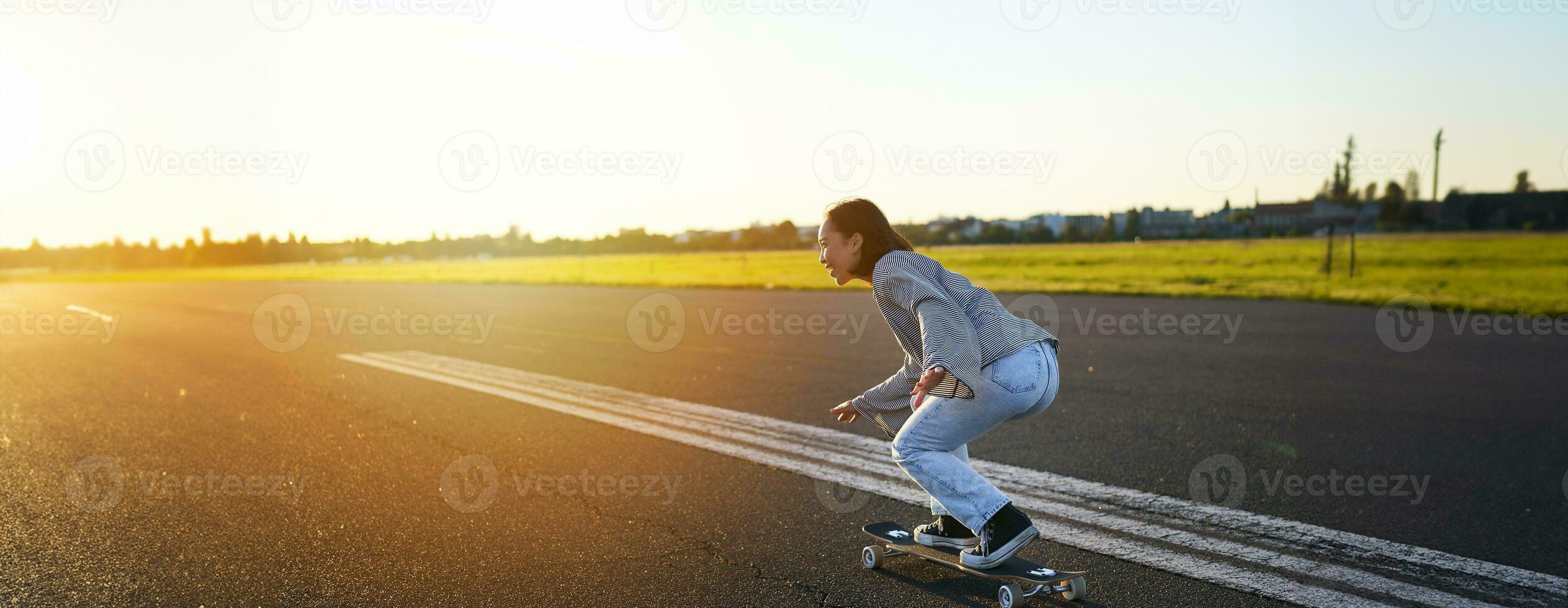 Side view of beautiful asian girl on skateboard, riding her cruiser towards the sun on an empty road. Happy young skater enjoying sunny day on her skate photo