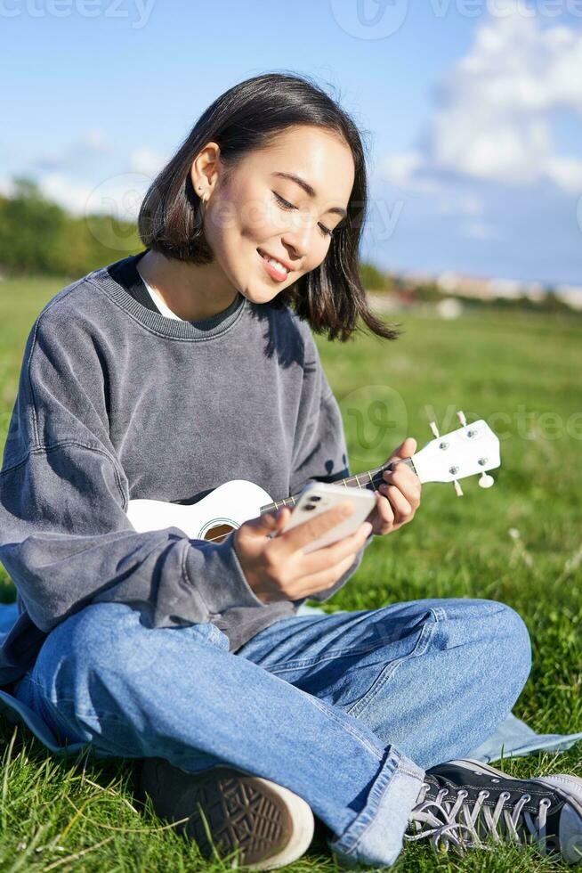 Vertical shot of smiling asian girl with smartphone, playing ukulele, reading chords or lyrics while singing, relaxing outdoors. Lifestyle and people concept photo