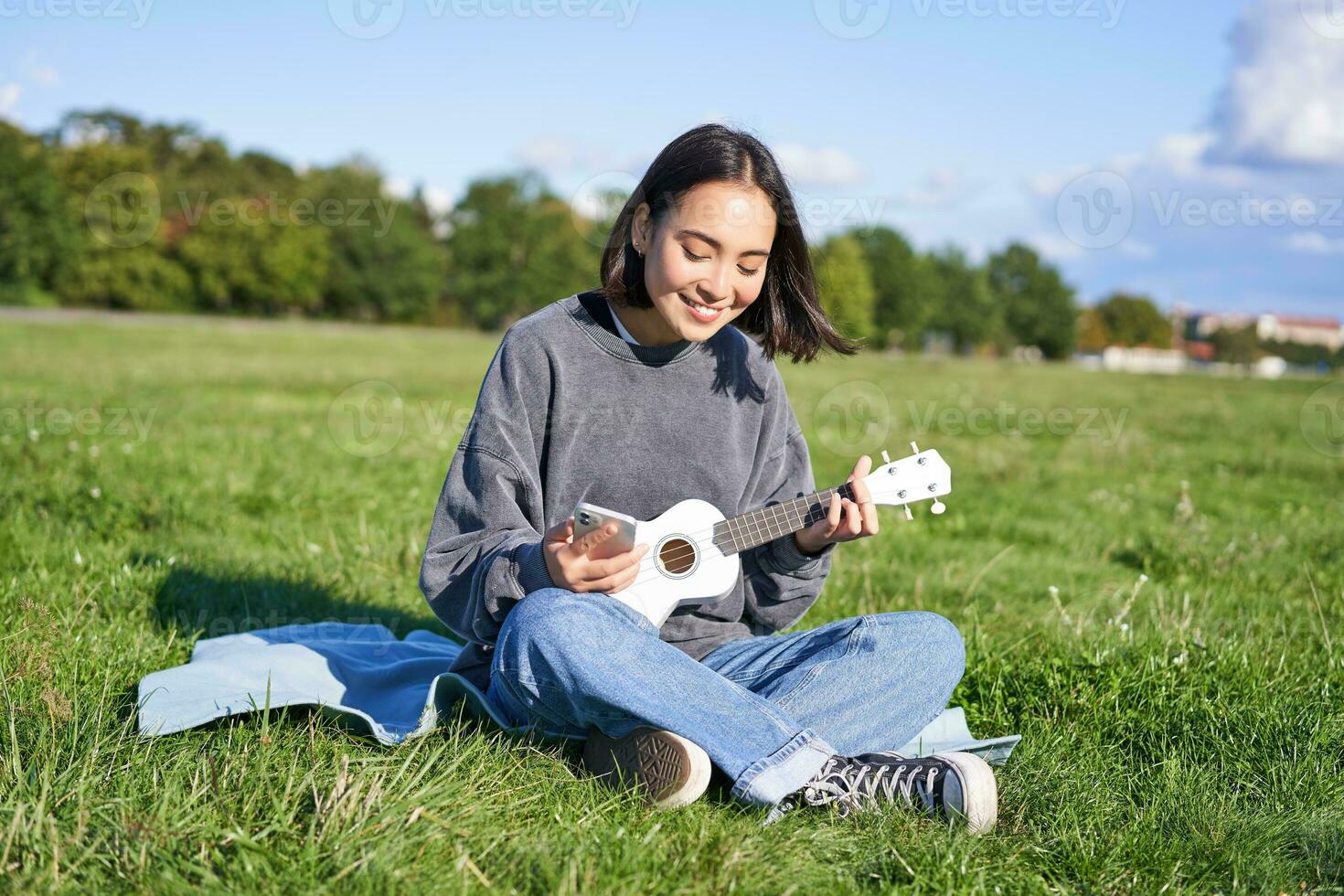 sonriente asiático niña mirando a guitarra aplicación, canto y jugando ukelele mientras echar un vistazo a acordes en teléfono inteligente, sentado al aire libre en parque en cobija foto