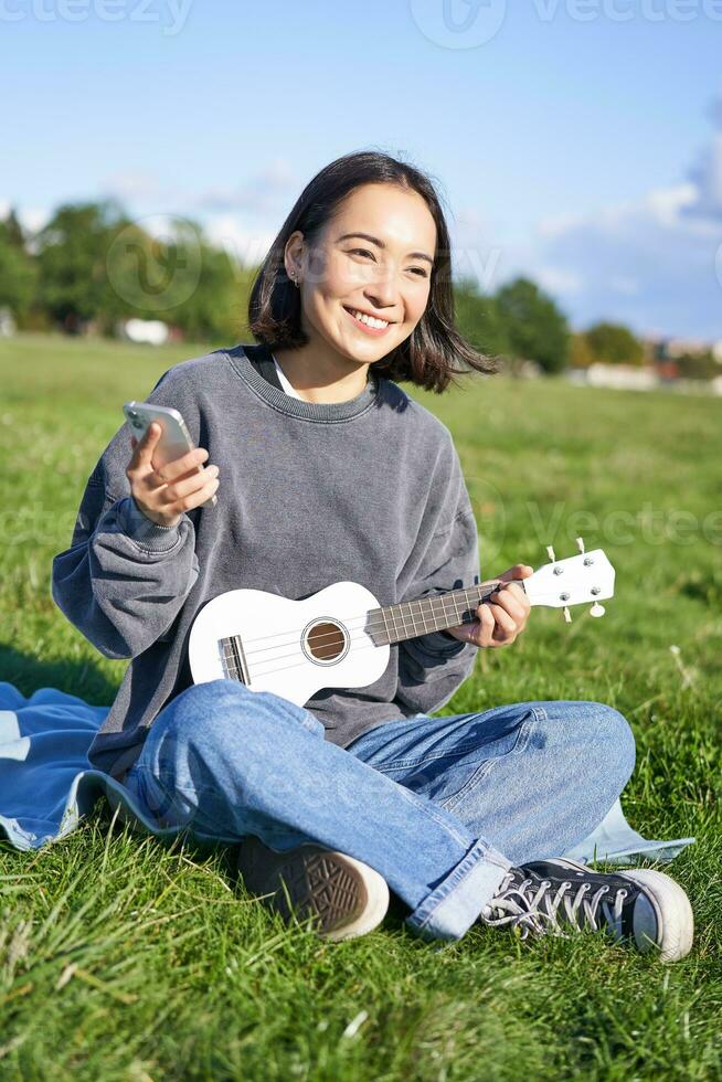 Vertical shot of smiling korean girl sitting in park with ukulele and smartphone, using mobile app to tune instrument, looking for chords while playing photo