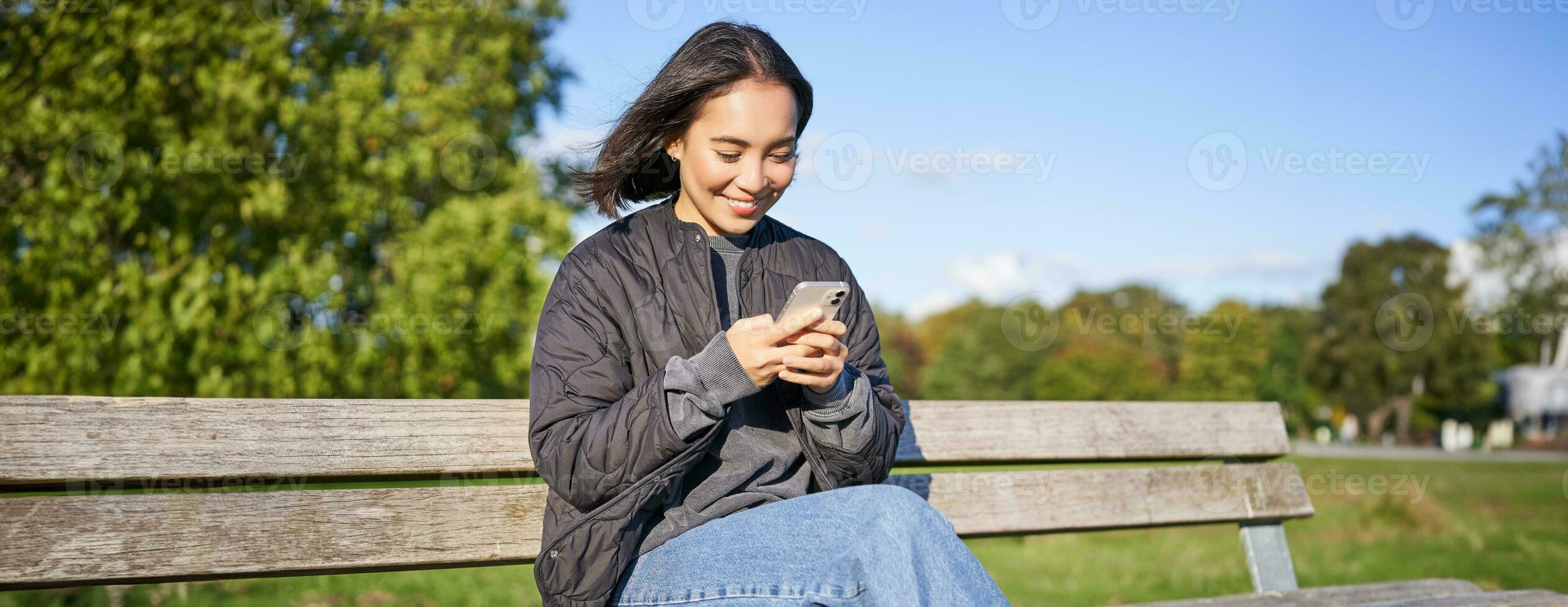 Technology and people. Young woman using smartphone, sitting outdoors on bench in park, looking at smartphone, sending message or networking photo