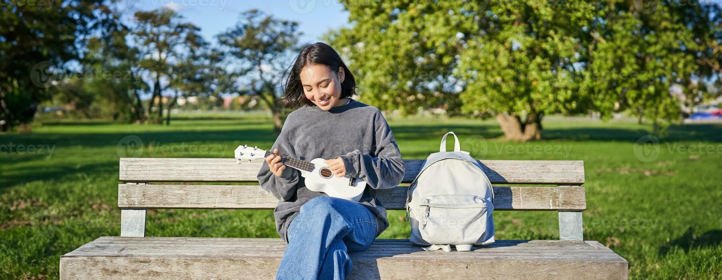hermosa asiático niña obras de teatro ukelele al aire libre, se sienta en parque en banco con musical instrumento foto
