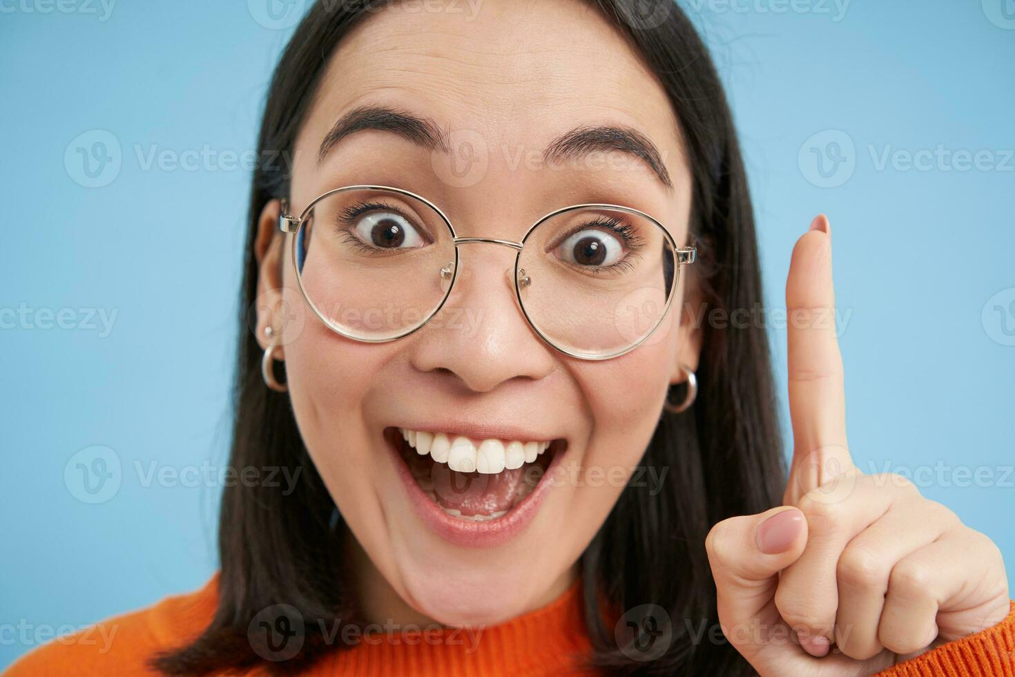 Close up portrait of happy asian girl in glasses, raises finger, has revelation, suggest great idea, says eureka, stands over blue background photo