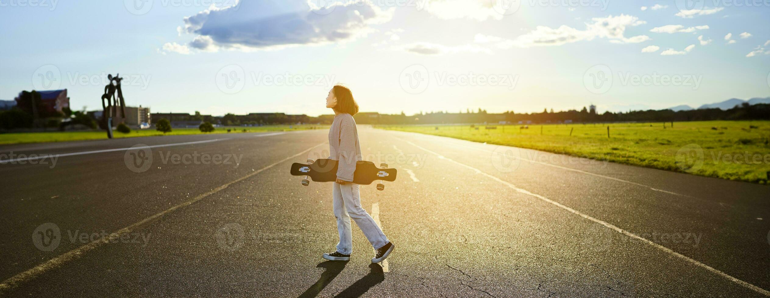 Young skater girl, teenager skating on cruiser, holding longboard and walking on concrete empty road photo