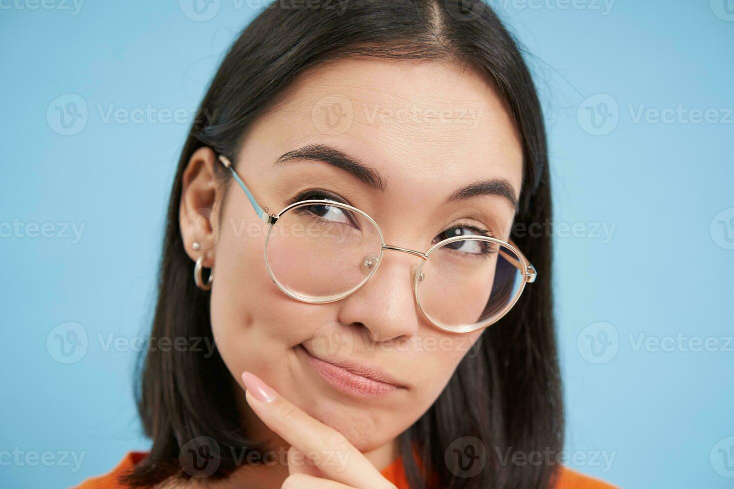 Portrait of Japanese woman in glasses, looks thoughtful, ponders, thinking with serious face, standing over blue background photo