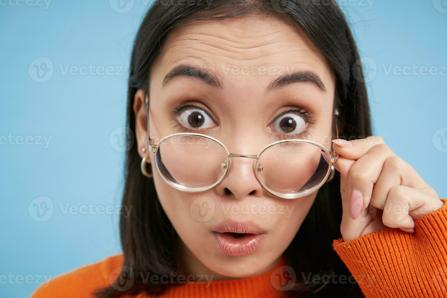 Portrait of amazed japanese woman in glasses, reads smth interesting, wears prescribed eyewear, blue background photo