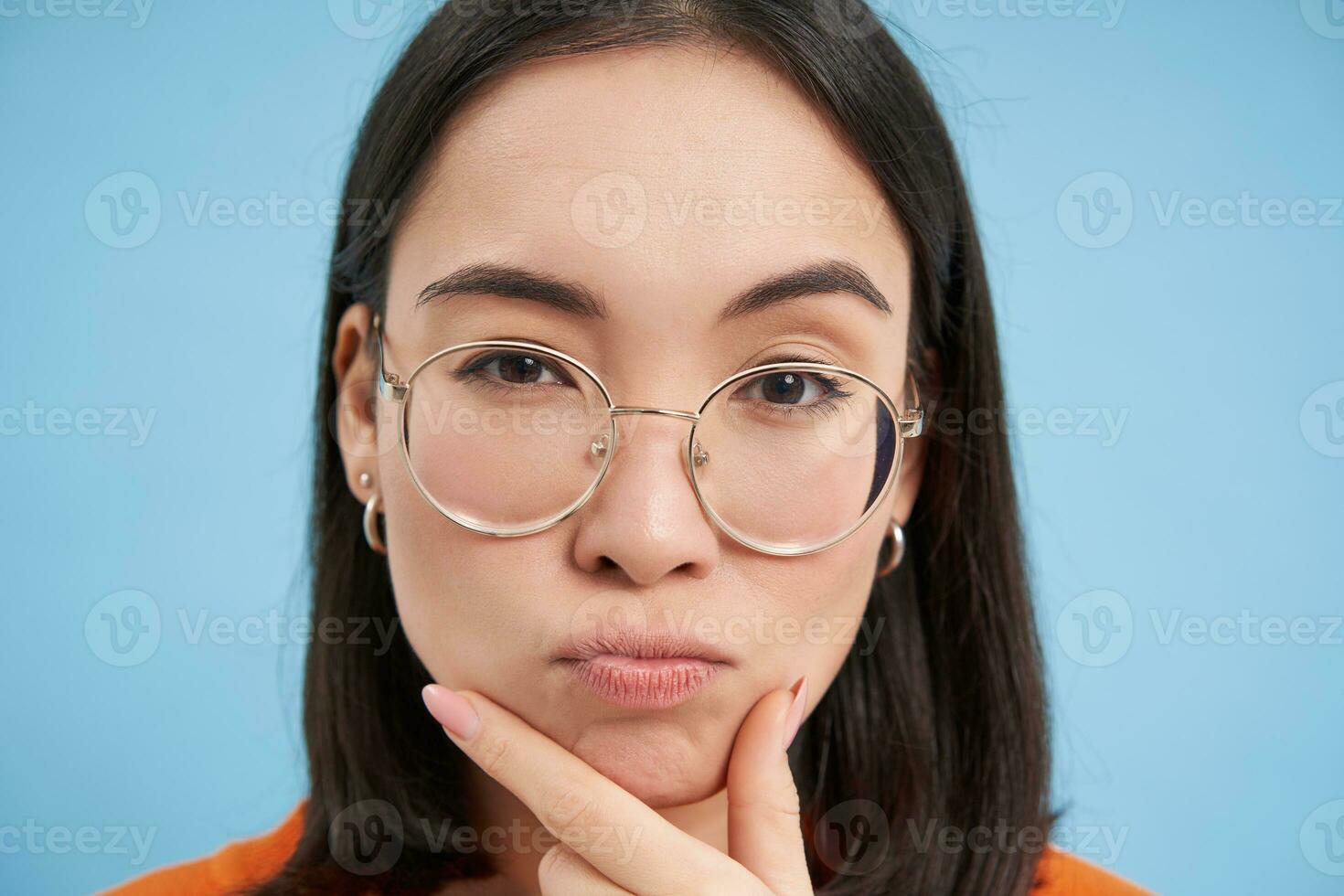 Close up portrait of asian girl in glasses, squints with suspicious face, thinking, stands over blue background photo