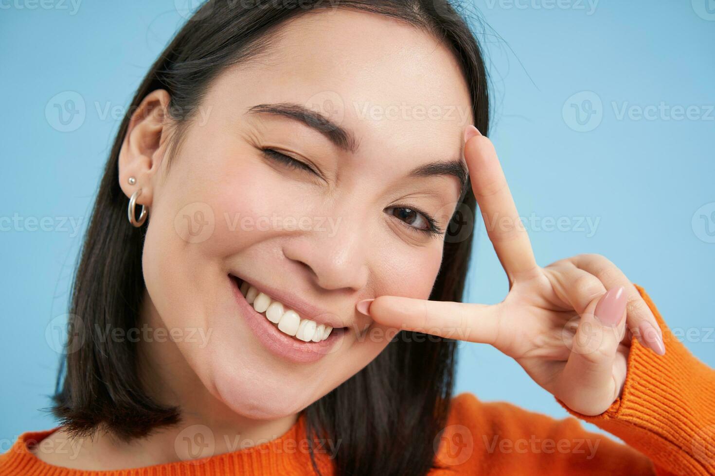 Close up portrait of positive asian woman, winks and shows peave, v-sign gesture, sticks tongue and smiles, stands over blue background photo