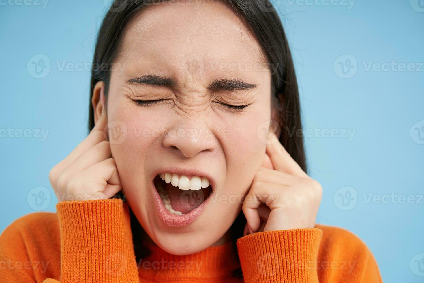 Close up portrait of screaming woman, shuts her ears with fingers and shouts, stands over blue background photo