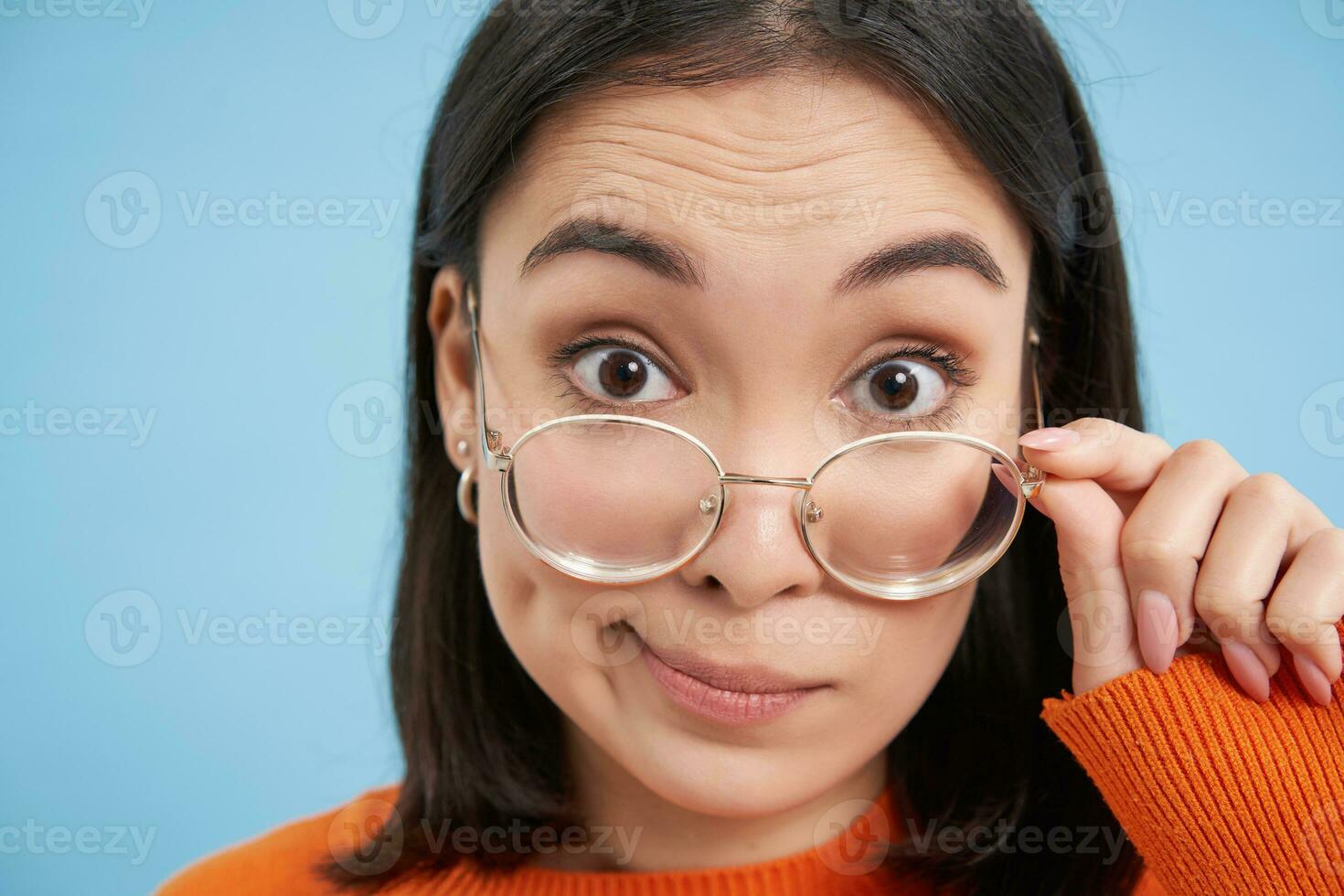 Close up portrait of amazed girl, looks closer at camera in glasses, standing against blue background photo