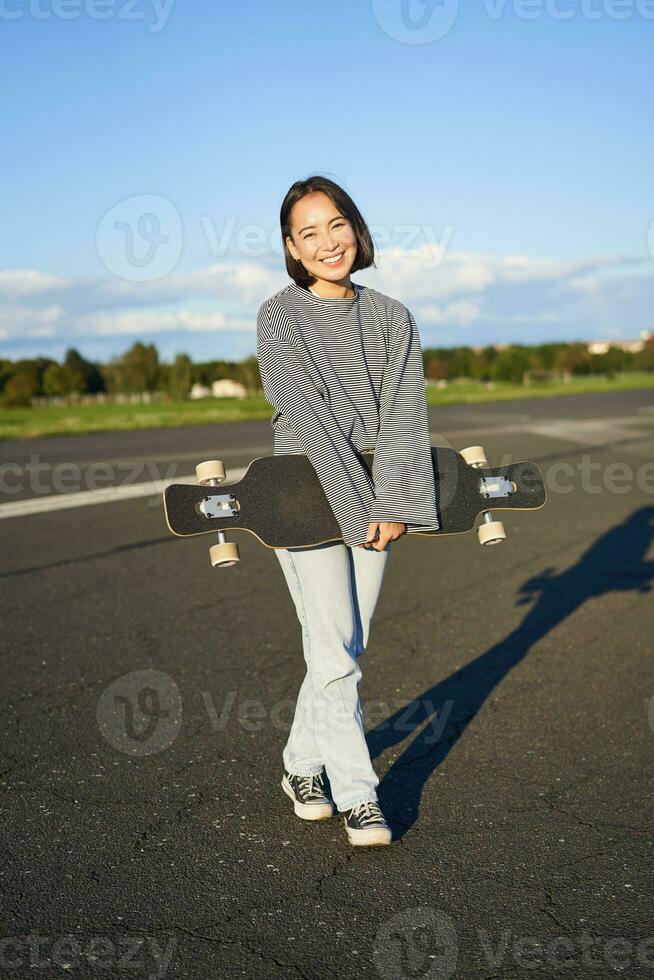 vertical Disparo de patinador niña posando con longboard, crucero en vacío la carretera en afueras. sonriente asiático mujer Patinaje en patineta, participación crucero en manos foto