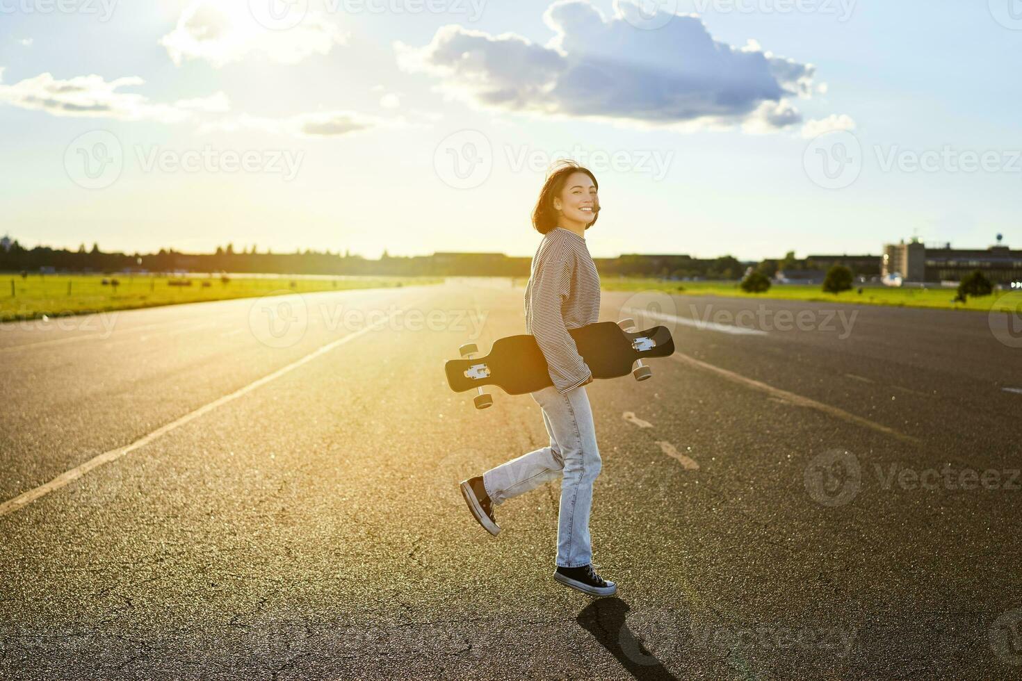 Asian girl with skateboard standing on road during sunset. Skater posing with her long board, cruiser deck during training photo
