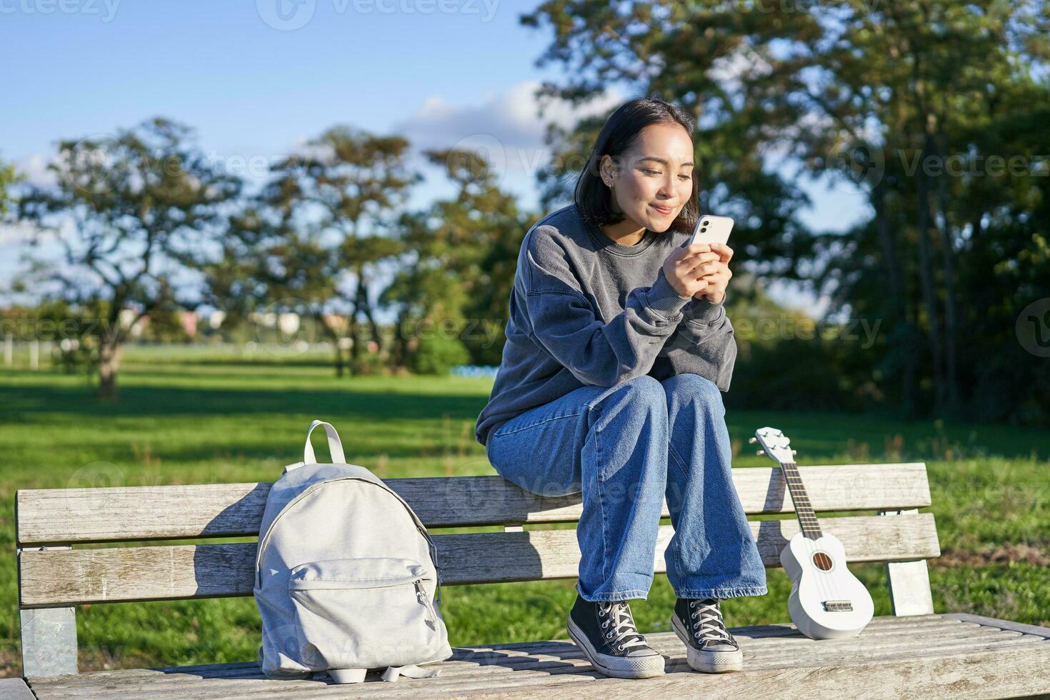 joven mujer sentado en parque en banco con ukelele, mirando a teléfono inteligente, leyendo mensaje en móvil teléfono y sonriente foto