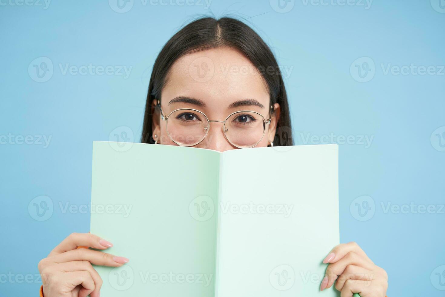 Portrait of cute japanese woman in glasses, peeking from notebook, covers face with her notes, smiles with eyes, blue background photo