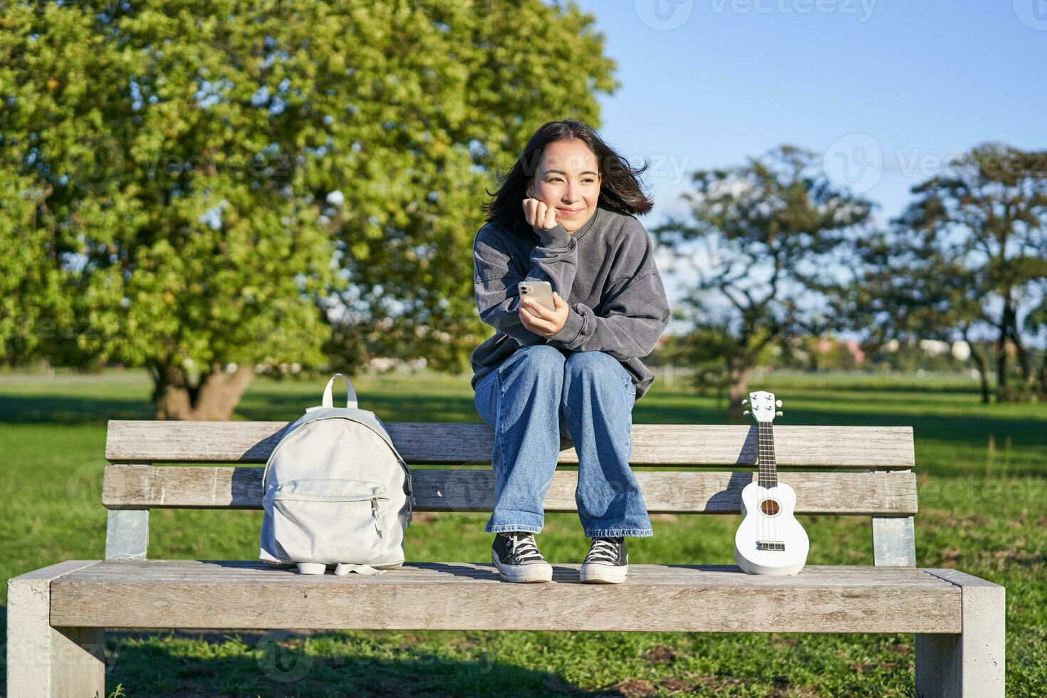 joven mujer con ukelele, sentado en banco en parque, utilizando móvil teléfono aplicación, participación teléfono inteligente y sonriente foto