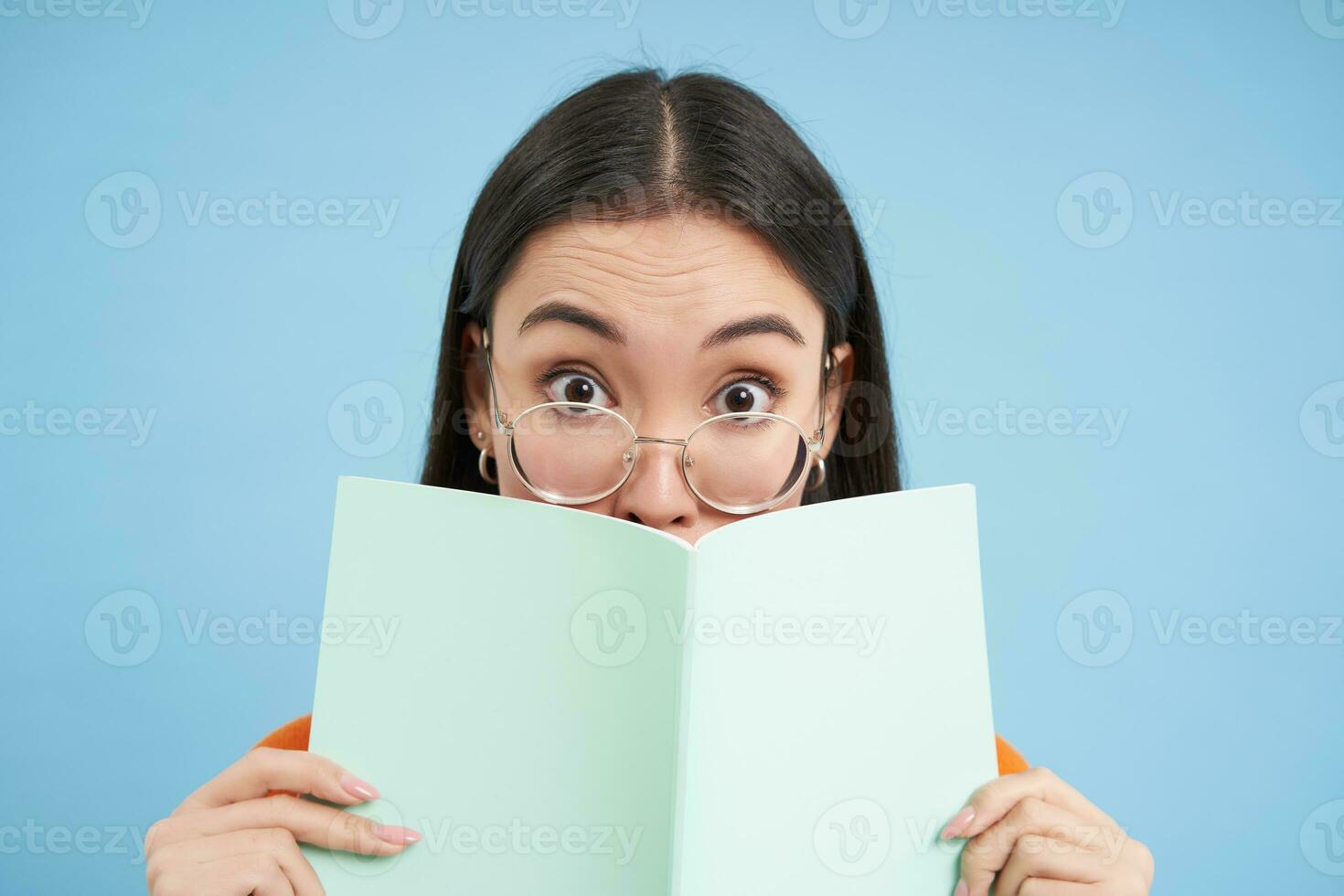 Portrait of cute japanese woman in glasses, peeking from notebook, covers face with her notes, smiles with eyes, blue background photo