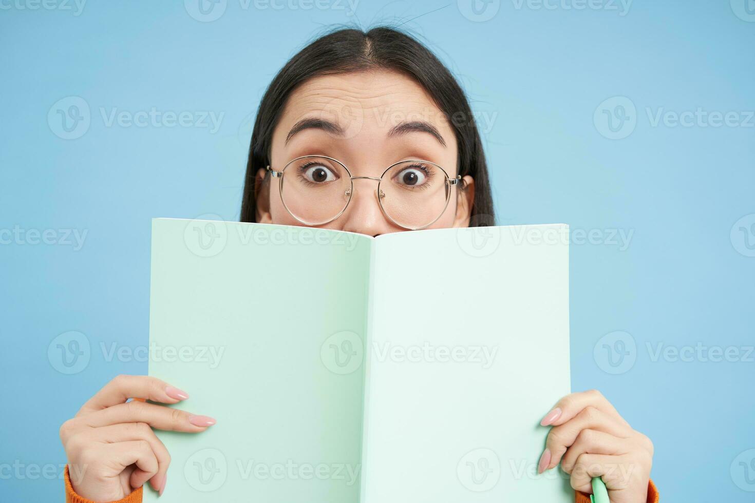 Cute asian woman in glasses, hides her face behind notebook and smiles with eyes, curious gaze, stands over blue background. Students and education concept photo