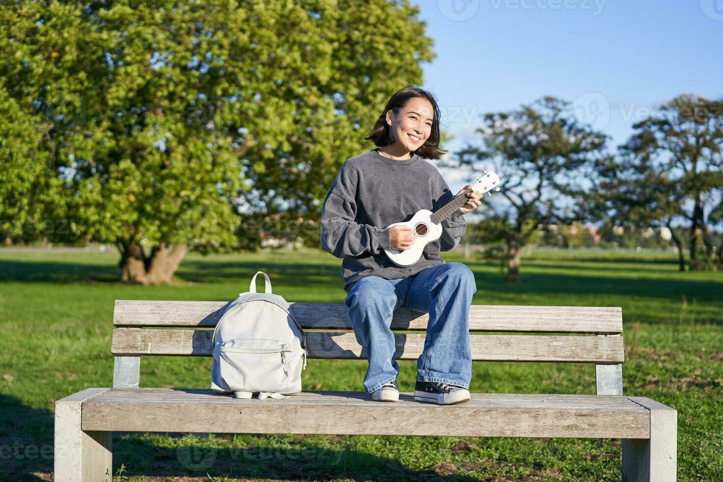 retrato de joven hipster niña se sienta en banco y obras de teatro ukelele, canta a lo largo de, relajante en parque con su musical instrumento foto