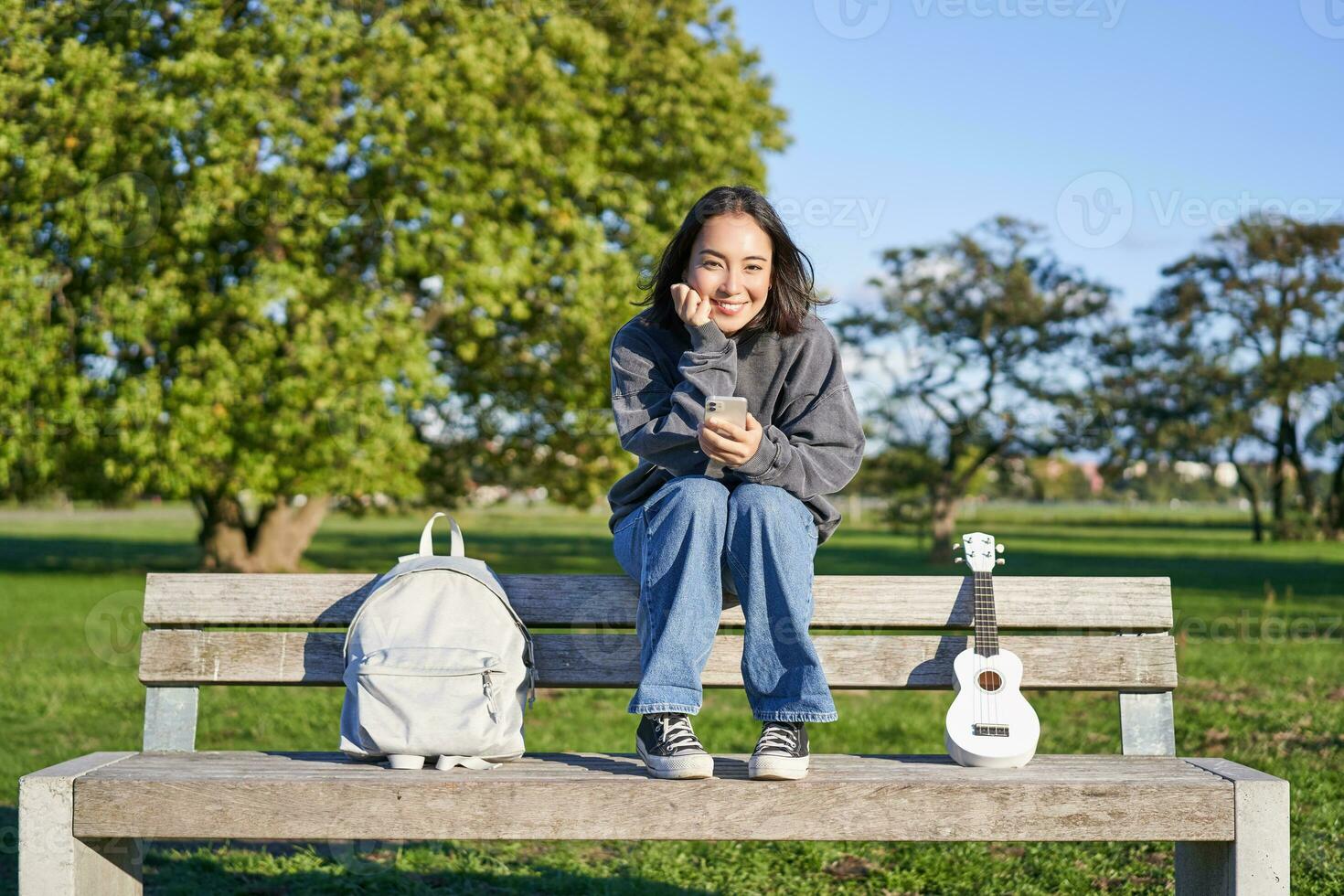 hermosa morena niña en banco en parque, sentado con ukelele y mochila, participación teléfono inteligente, utilizando móvil aplicación foto