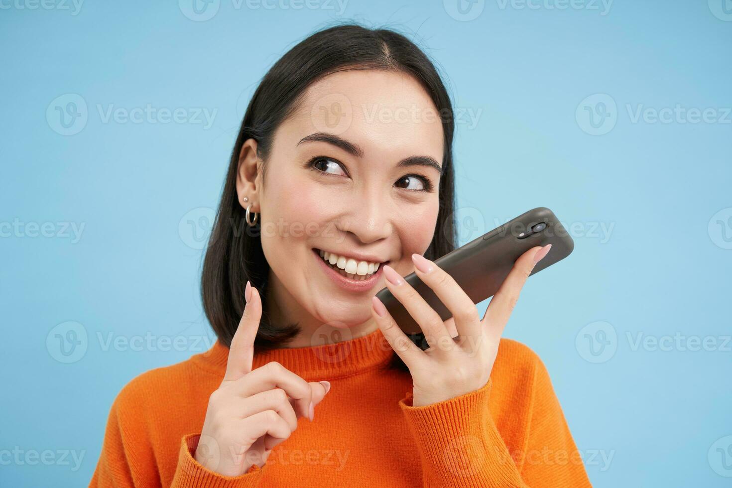 Portrait of korean woman records voice message on smartphone app, talks on speaker, translates her speech on mobile app, blue background photo