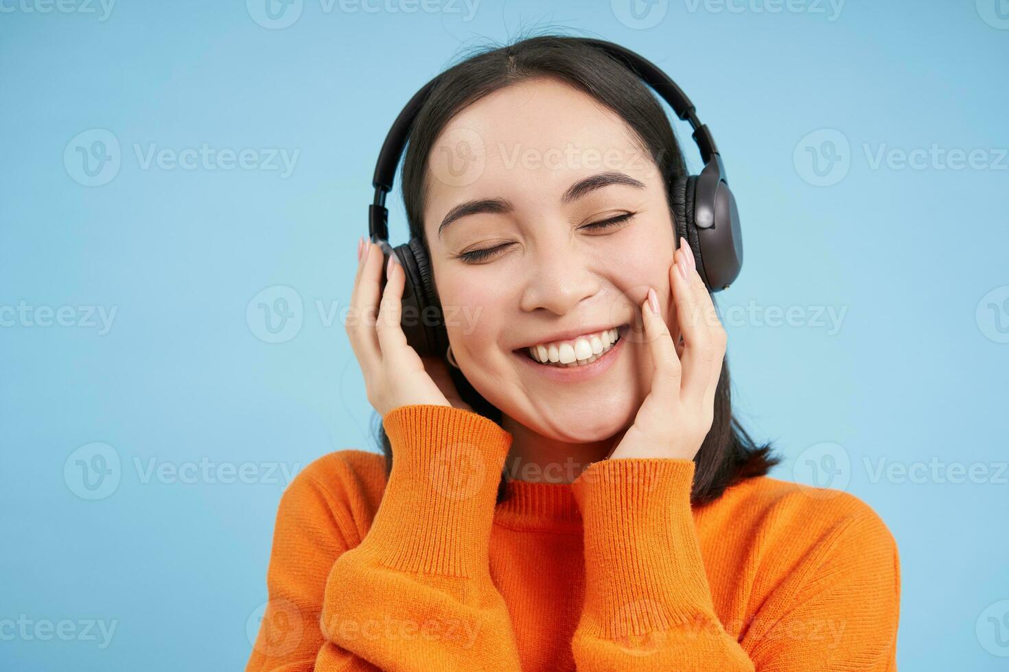 Close up of beautiful asian woman in headphones, smiles and listens music, enjoys podcast, stands over blue background photo