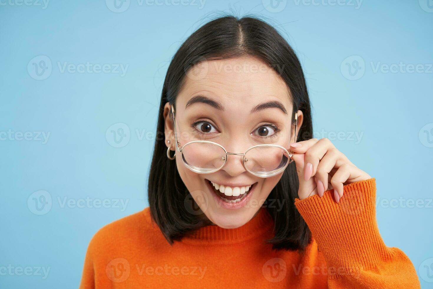 Portrait of japanese girl, takes of glasses, stares at camera in eyewear, looks amazed, blue background photo