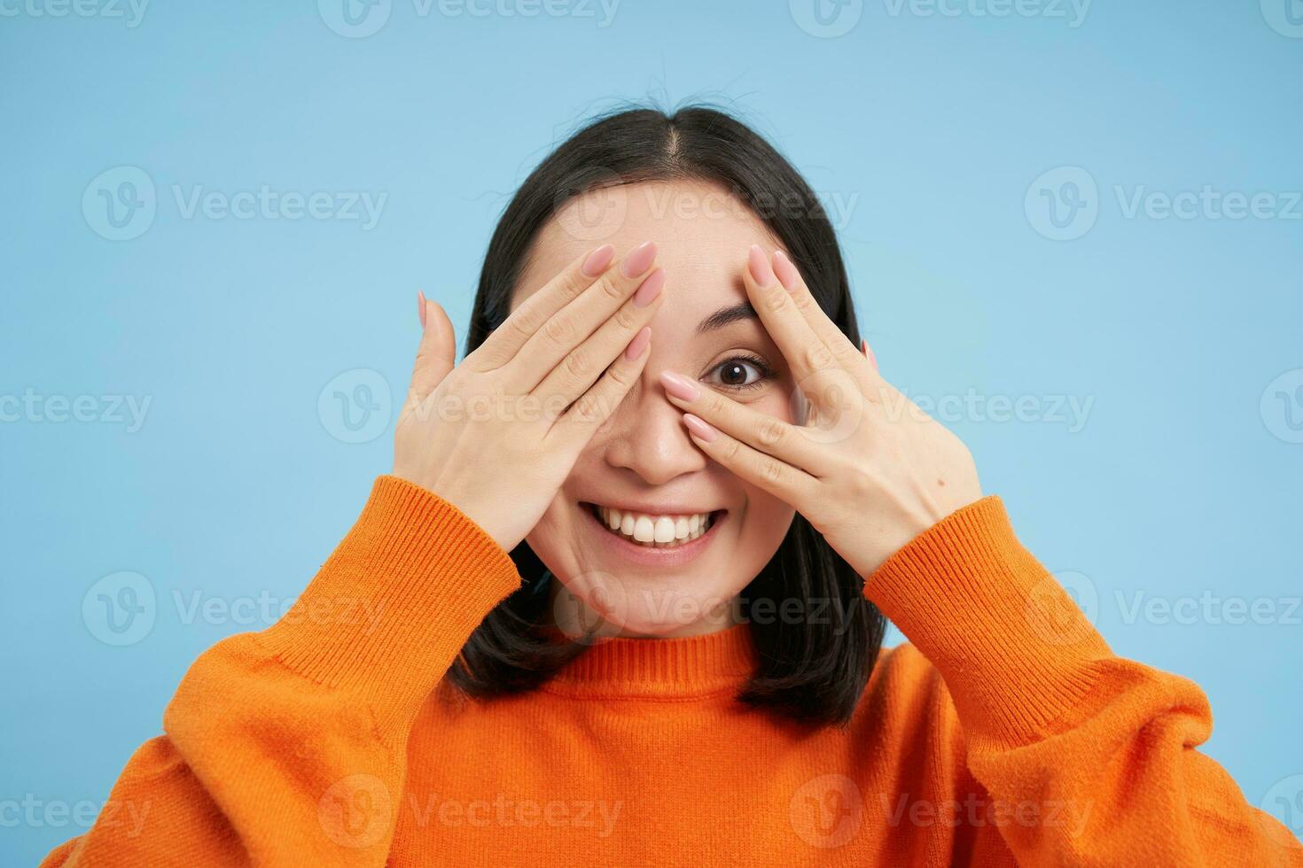 Close up portrait of korean girl waits for surprise, shuts her eyes and peeks through fingers with excited smile, stands over blue background photo