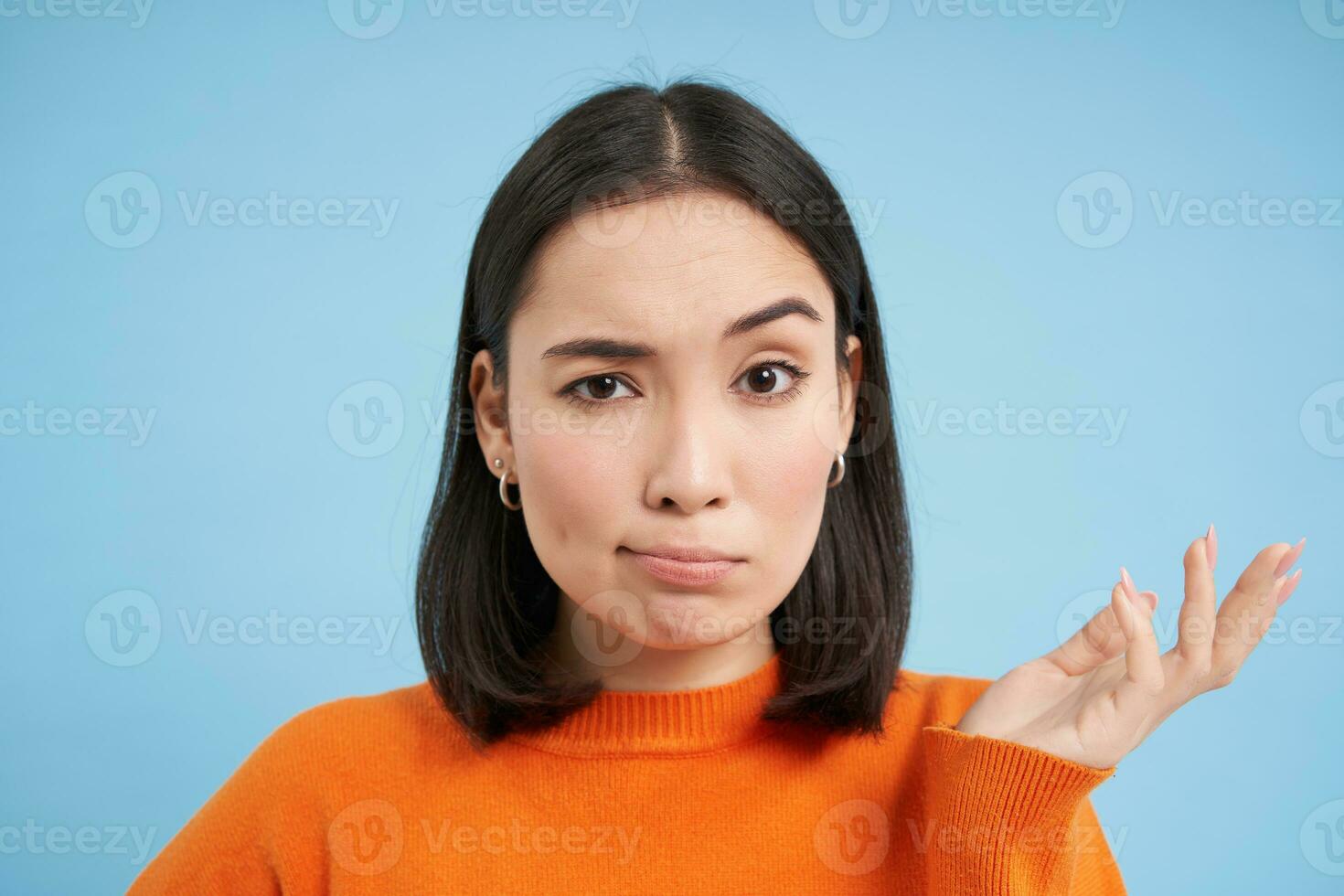 So what. Portrait of korean woman shrugs and raises arm up with skeptical, confused face expression, stands over blue background photo